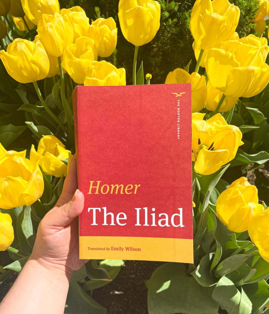 Red and yellow cover of Emily Wilson’s translation of Homer’s The Iliad for the Norton Library series against a backdrop of yellow tulips. 