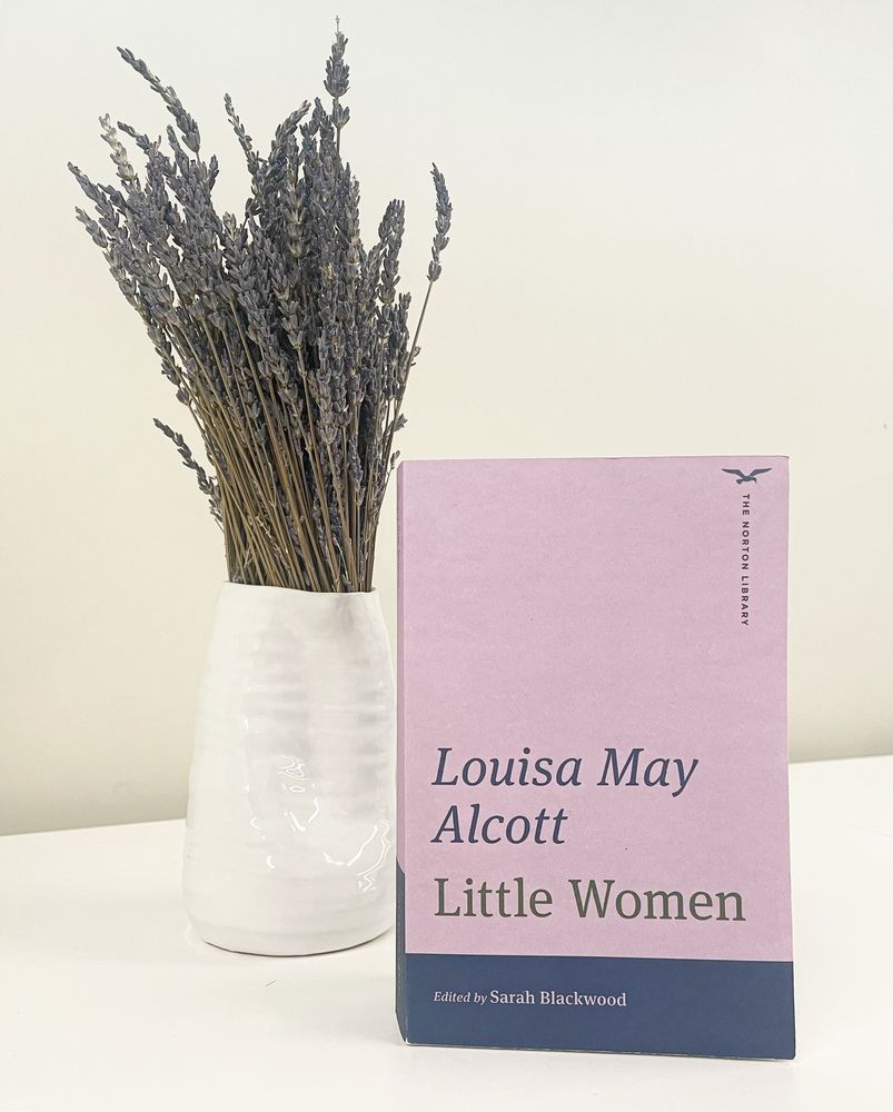 Norton library edition of Louisa May Alcott’s Little Women. The purple book is against a white background with a white vase of dried lavender sprigs 