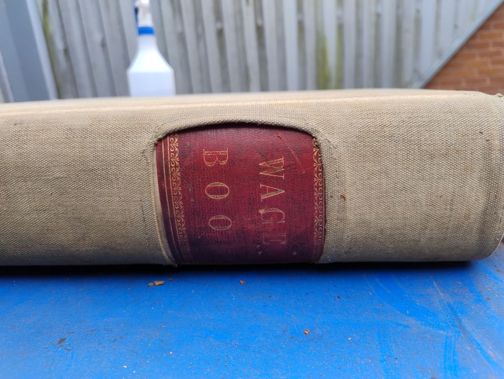 Archive volume lying on its side on a document trolley. The canvas book jacket has a cutaway that allows the gold tooled wording "WAGES BOOK" to be seen.