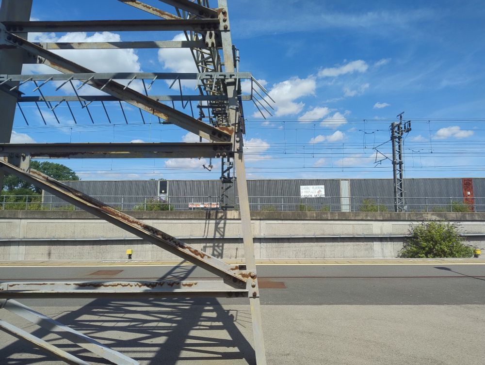 View from a train in Nuneaton Station. A metal tower similar to a pylon is on the left of the image. A gantry is to the right. A sign on the corrugated steel wall in the background reads "Classic flooring and fireplaces".