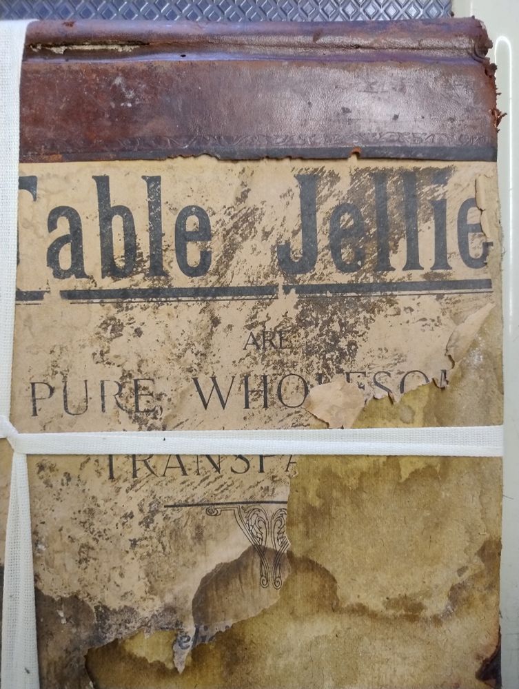 Close up of the back of a leather and book cloth bound volume. The book cloth has disappeared from the back board at some point, revealing an advert for table jellies. The volume is held together with archival tape. The wording reads "Table Jellies are Pure, Wholesome and Transpa[rent]."
