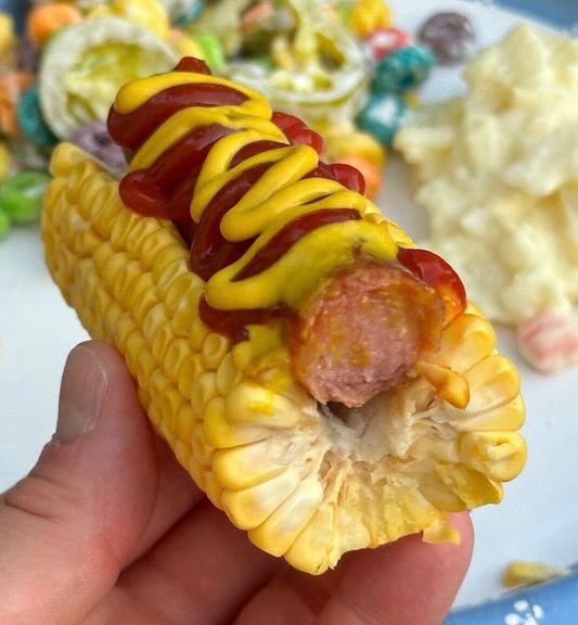 A cooked ear of corn with a length of it sliced out so that it can fit a hot dog as if the air of corn or a bun. There’s a hotdog in it, covered in ketchup and mustard.