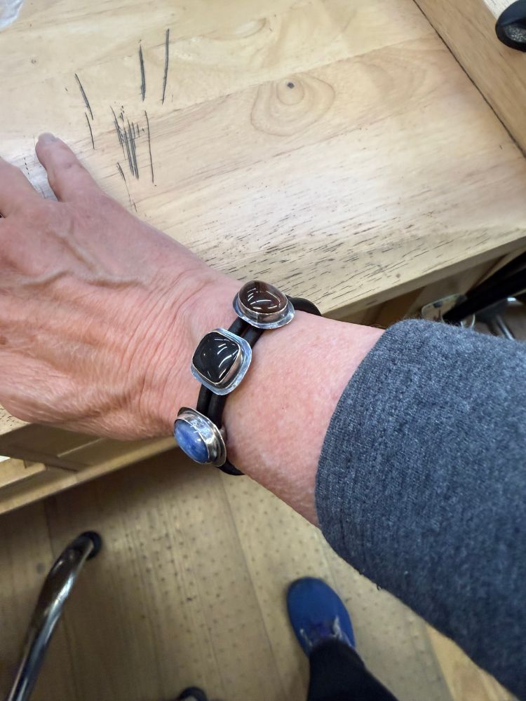 A woman’s wrist and arm resting on a jeweler’s bench, showing off a silver and black leather bracelet with kyanite, onyx, and agate set stones sliding on the leather. 