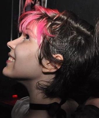 Foto colorida de uma pessoa de pele bege de lado, sorrindo, olhando um tanto pra cima. Elu tem cabelo castanho bem curtinho, raspado dos lados e com uma franja rosa. O nariz é reto e um pouco arrebitado.