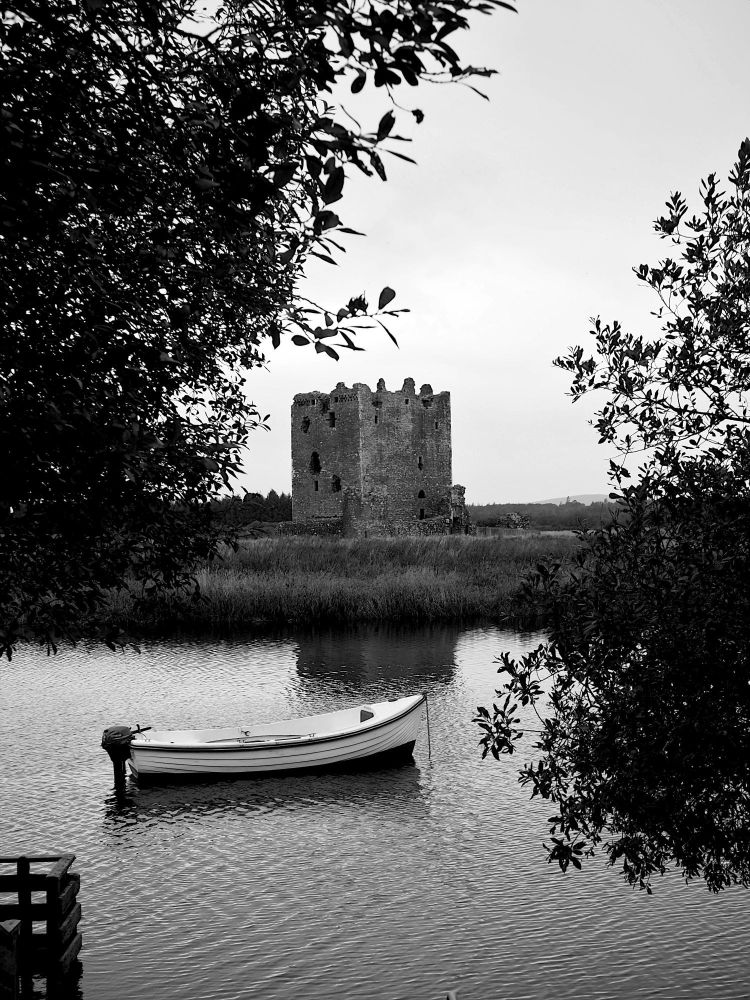 Black and white view of an empty small boat in the River Dee, framed by trees in the foreground, with the square keep of Threave Castle and a featureless sky in the background.