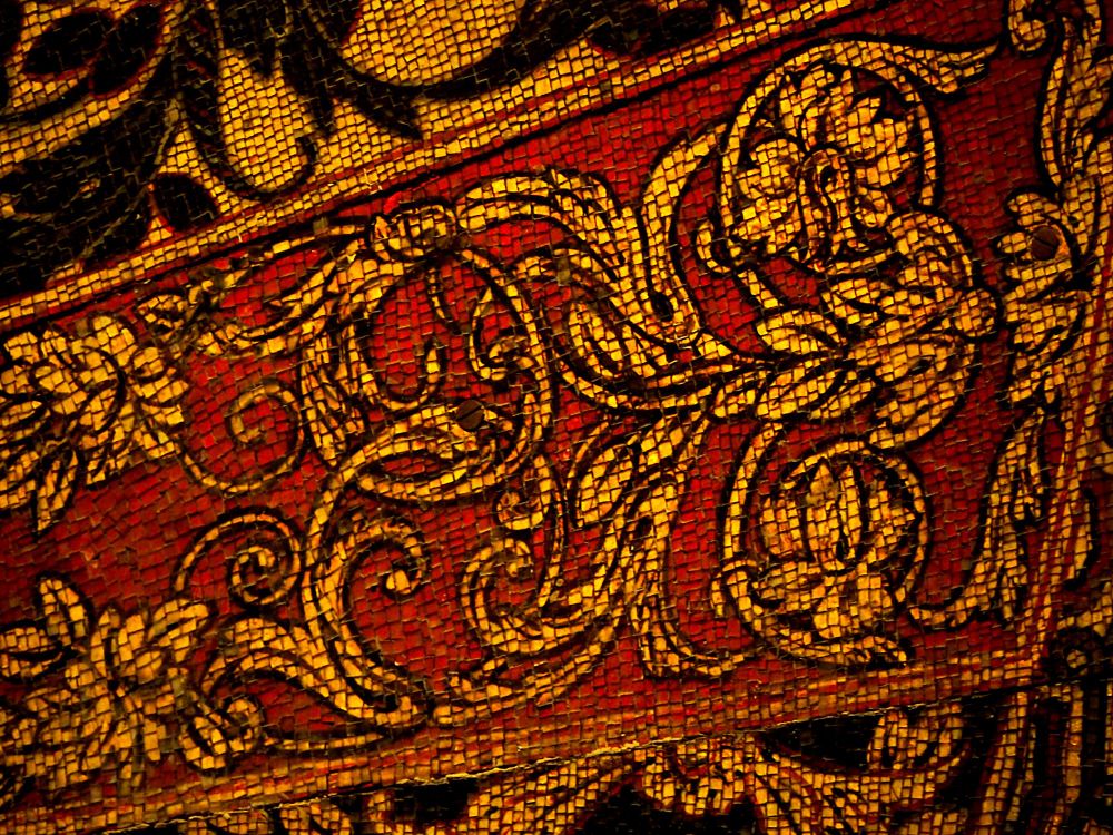 Detail photograph of a section of mosaic, mainly featuring entwined leafy tendrils in gold on a red background. At top-left and bottom-right, the mosaic continues in black and gold.