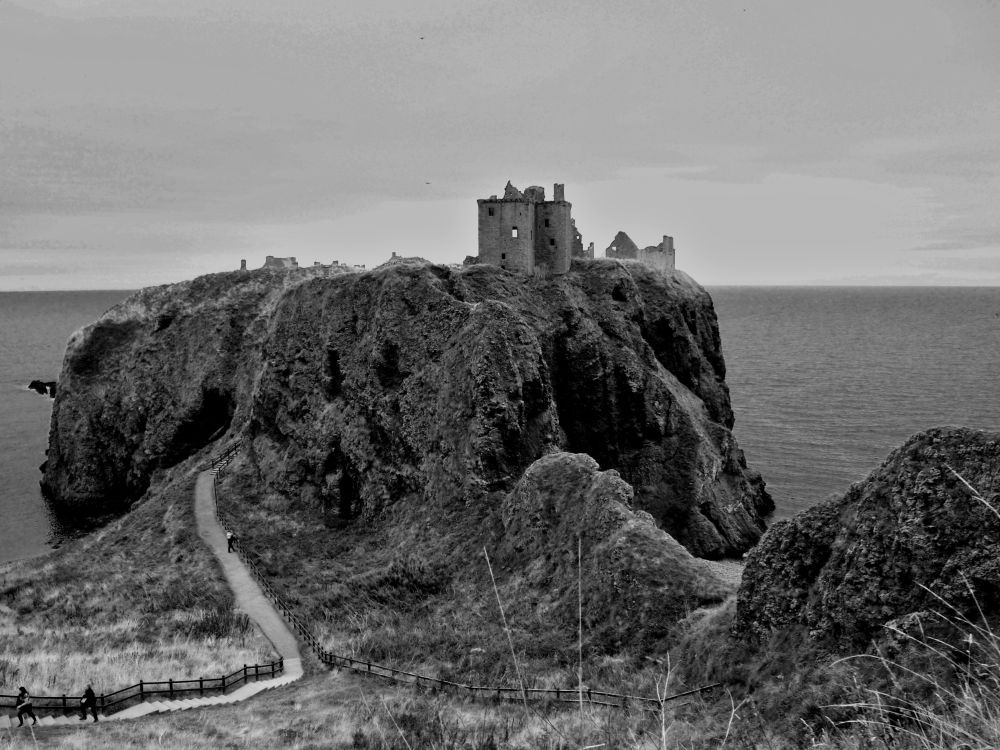Black and white image of ruined castle buildings perched on a rocky outcrop in centre of frame, with sea and sky beyond and either side. A path leads toward the castle from the left foreground.