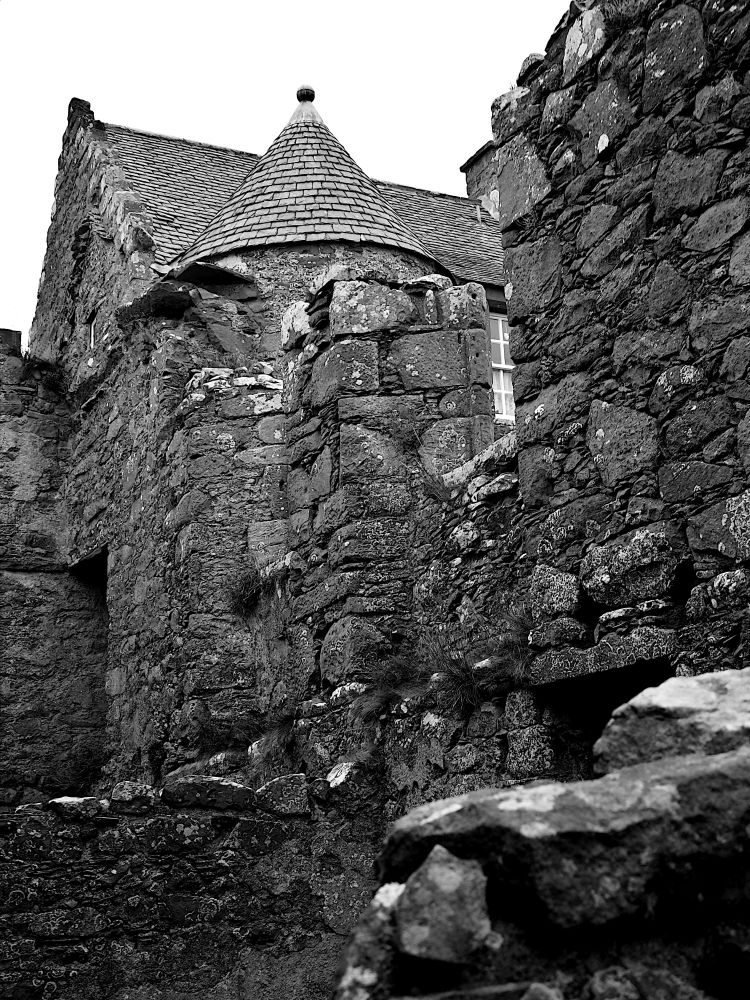 Black and white image of ruinous stone walls in foreground, looking up to a conically roofed circular turret and gabled building beyond.