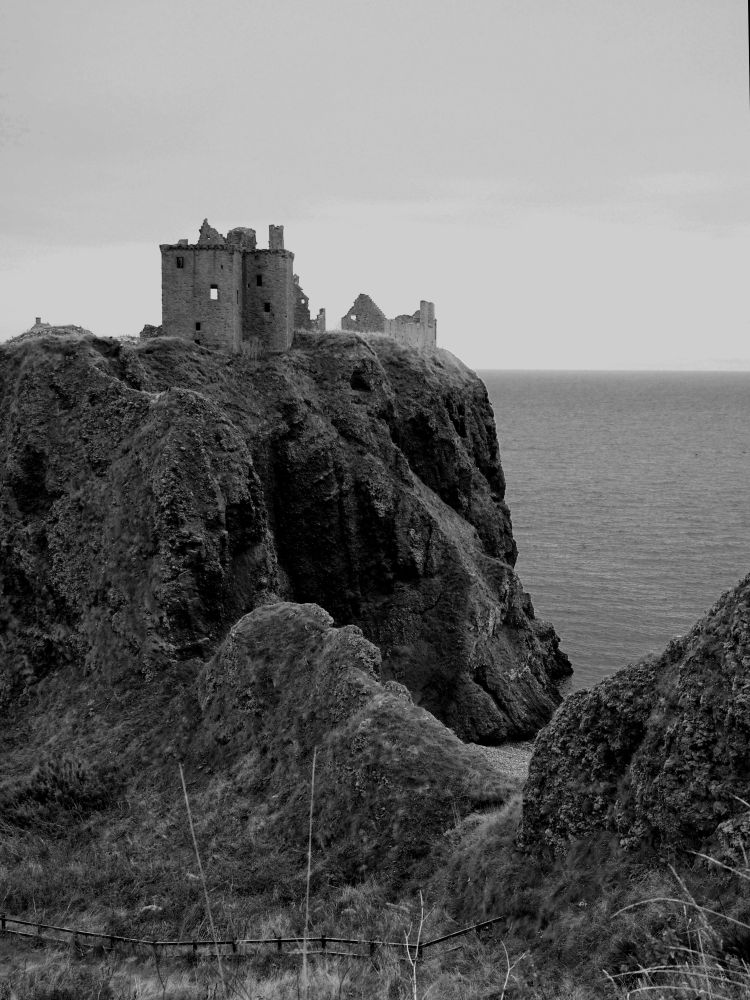 Black and white image of ruined castle buildings perched on a rocky outcrop toward the left of frame, with sea and sky beyond and to the right. Rugged terrain in foreground.