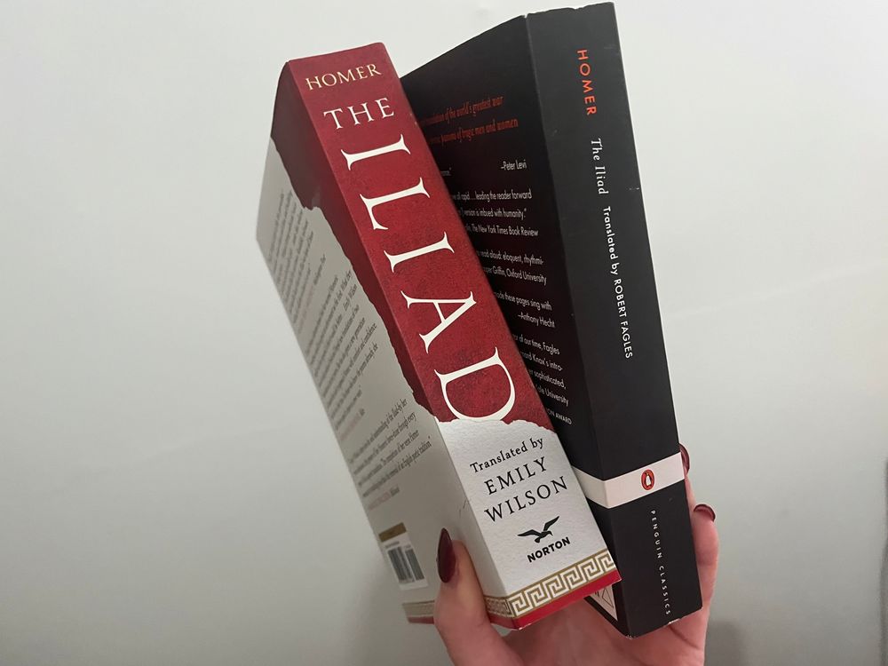 A Caucasian hand holding two books so their spines are visible. One is red and white and the other is black. Both are copies of the Iliad, the former translated by Emily Wilson, the latter by Robert Fagles