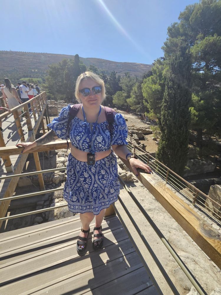 A blonde Caucasian woman wearing a blue and white crop top and skirt on a balcony. Behind her are ruins and trees 