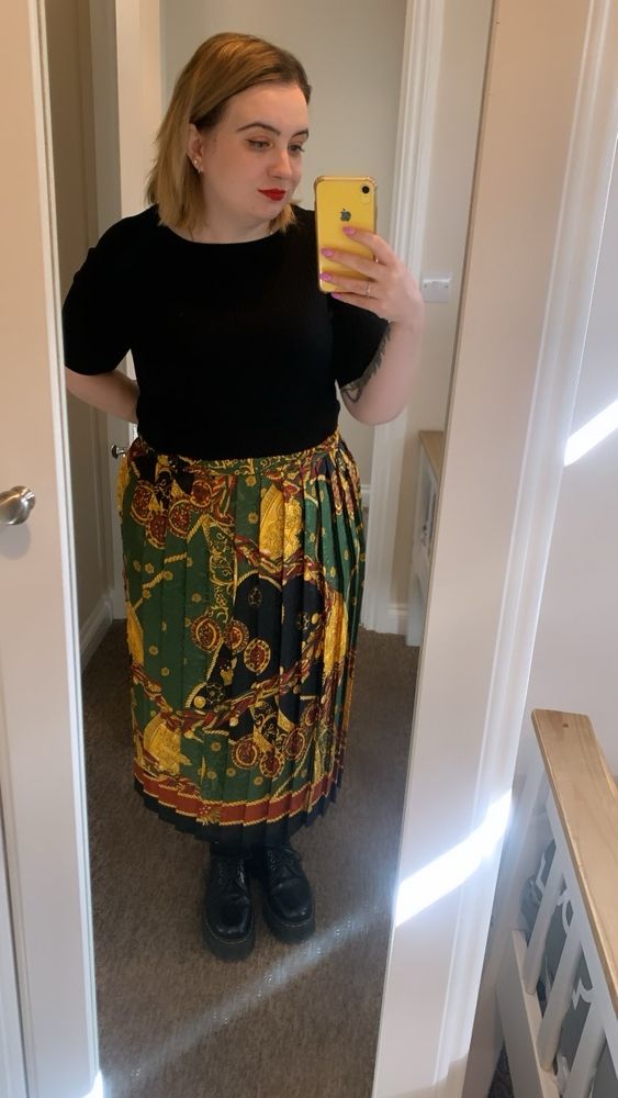 A white woman with shoulder length dark blonde hair taking a selfie in a mirror. She is wearing a black t shirt and a long, pleated black, gold, and green skirt