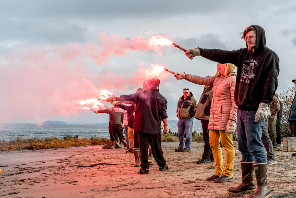 A group of people stand in a line on the beach holding flairs. 