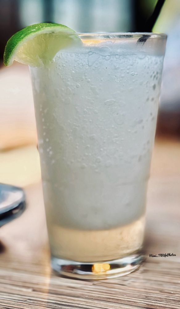 A glass sitting on a table in a restaurant with a frozen margarita and a lime hanging on the side of the glass with a straw sticking out of the top of the glass. 