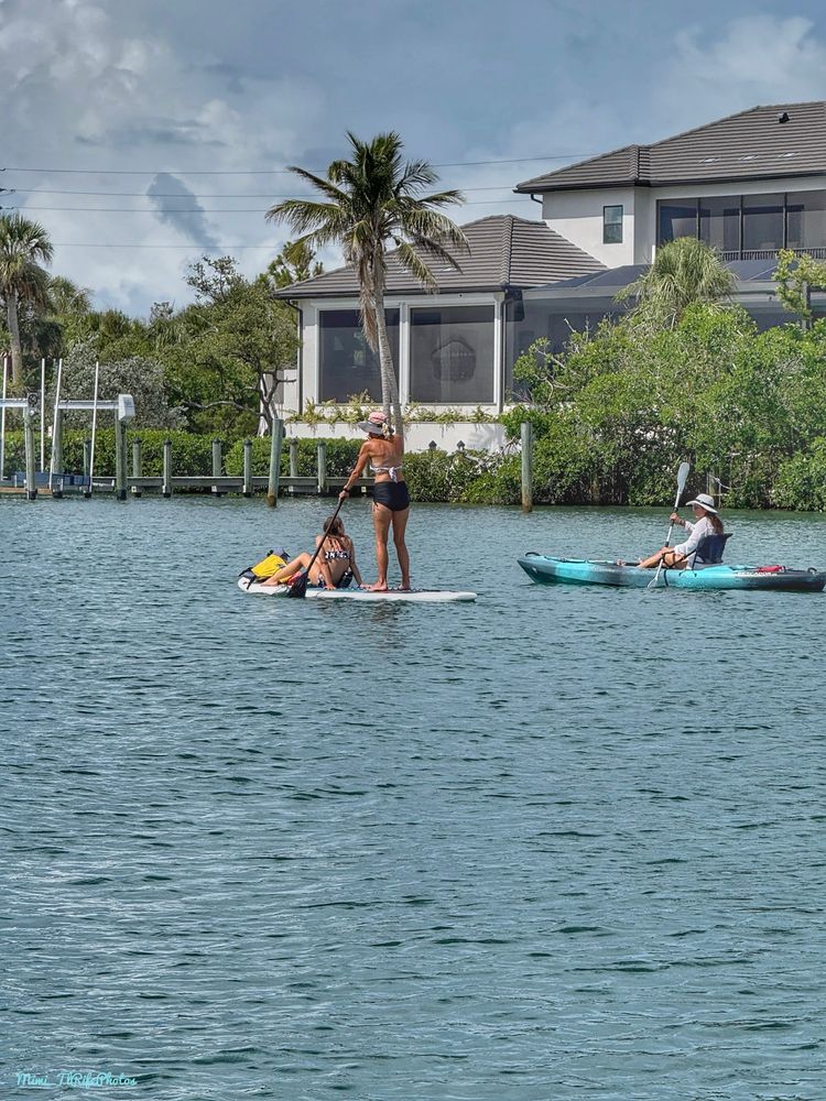 A young girl sitting on the front of a paddle board with a lady in a dark swim bottoms and a light top wearing a hat, rowing with a paddle. There is another person next to them in a light green and black kayak wearing a light colored shirt and colored hat.