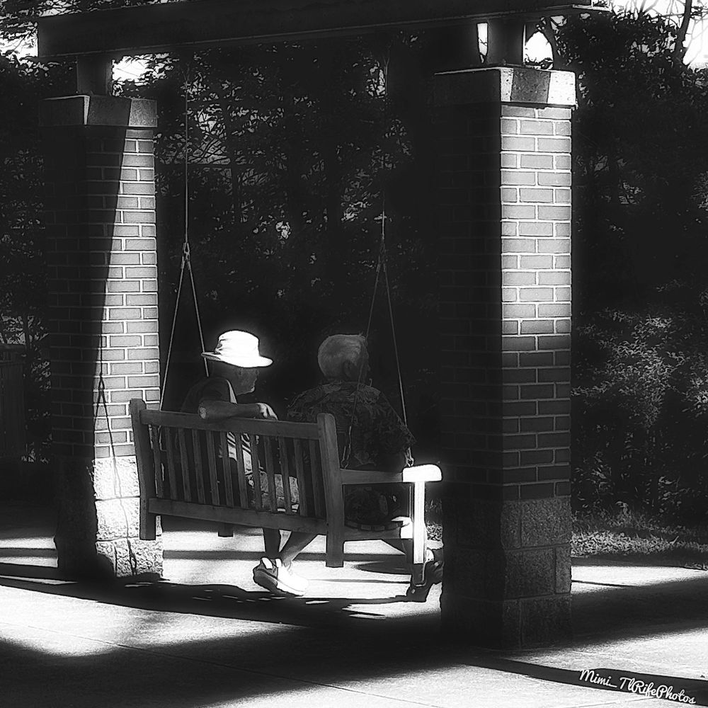 A black-and-white photo of two men sitting on a swing talking while enjoying the afternoon.