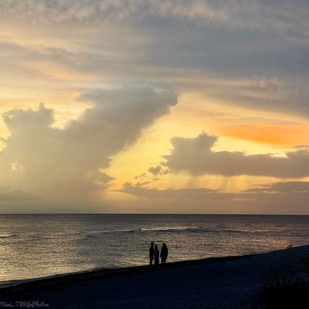 Three people are standing on the beach as the sun is setting with beautiful shades of orange, yellow, white, and peach. Golden Beach Venice, Florida 2025