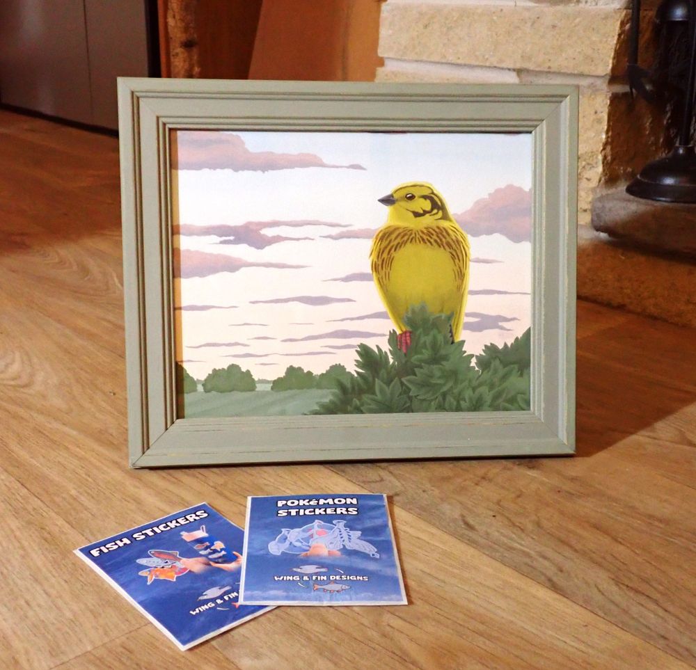 A photograph of a framed painting and a couple of sticker packs laid out on a wooden living room floor. The frame of the painting is a soft meadow green and the painting itself is of a Yellowhammer perched on a hawthorn hedge with crop fields in the background. There is a sunset sky with pink and purple clouds.
The sticker packs display the words "Fish stickers" and "Pokemon stickers".