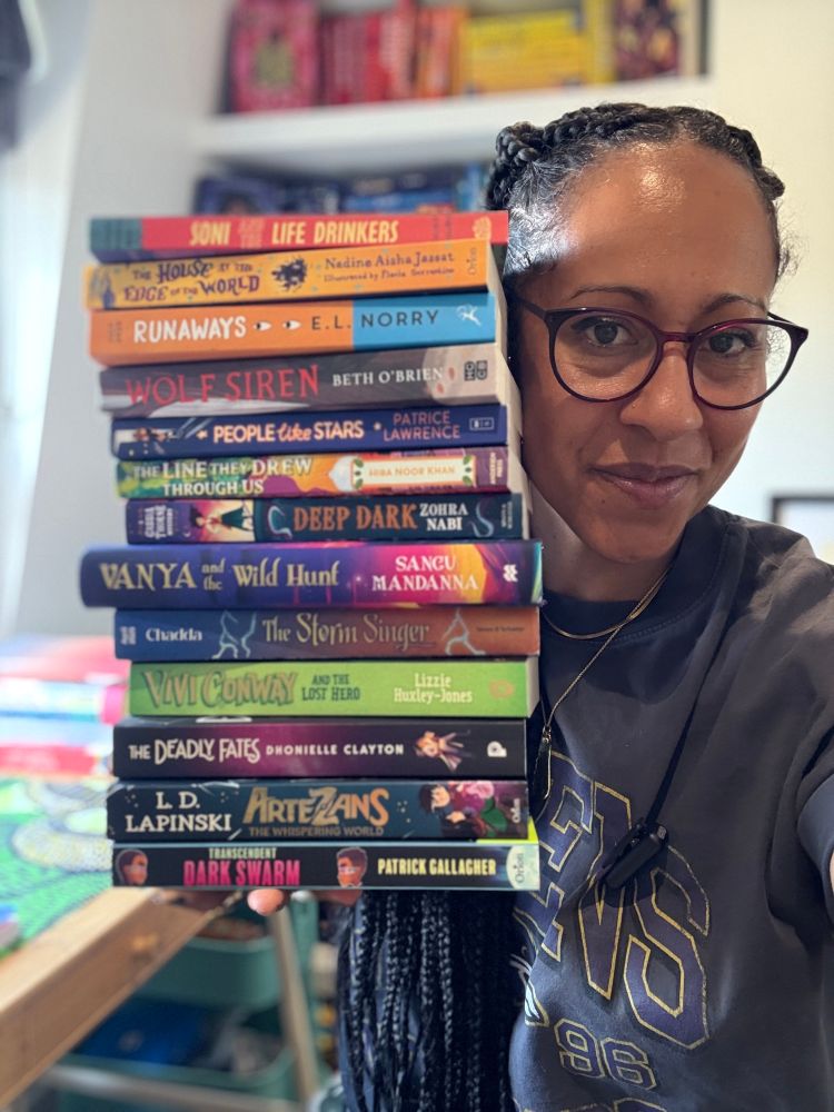 Claire, a mixed race woman holds a huge stack of middle grade books. 
Soni and the Life Drinkers 
Artezans: The Whispering World 
The Deadly Fates  
Vivi Conway and the Lost Hero by
The Runaways
The Line They Drew Through Us by   
People Like Stars 
Deep Dark 
The House at the Edge of the World
Wolf Siren  
Vanya and the Wild Hunt
The Storm Singer 