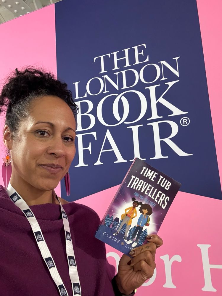 Claire, a mixed race woman with a pineapple bun standing in front of a London Book Fair sign holding a copy of Time Tub Travellers and the Silk Thief