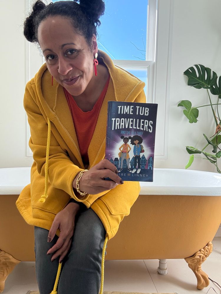 Claire sits on the edge of a yellow victors bath tub holding her book Time Tub Travellers. She has Afro puff buns and wears yellow and orange like the main character on the cover. 