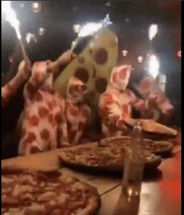 Image of people in pizza onesies, faces covered, waving sparklers at a table full of pizza pies
