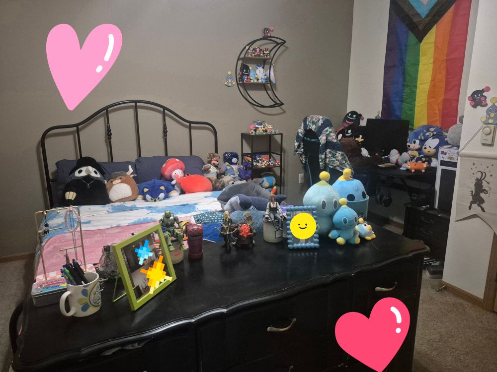 Bedroom with sky blue bedding with sonic plushes on top,there is a dresser that is short with nine drawers in front of the bed. To the side of the bed there is a small blue table and shelf holding various plush. Further to the side in a slight knock is a computer desk with gay flag above it. Desk is cluttered with nostalgic plushies.