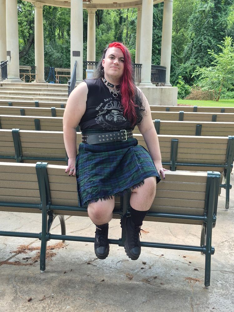 Plus sized goth man with long red and black hair with shaved sides. Black tank top, kilt and black