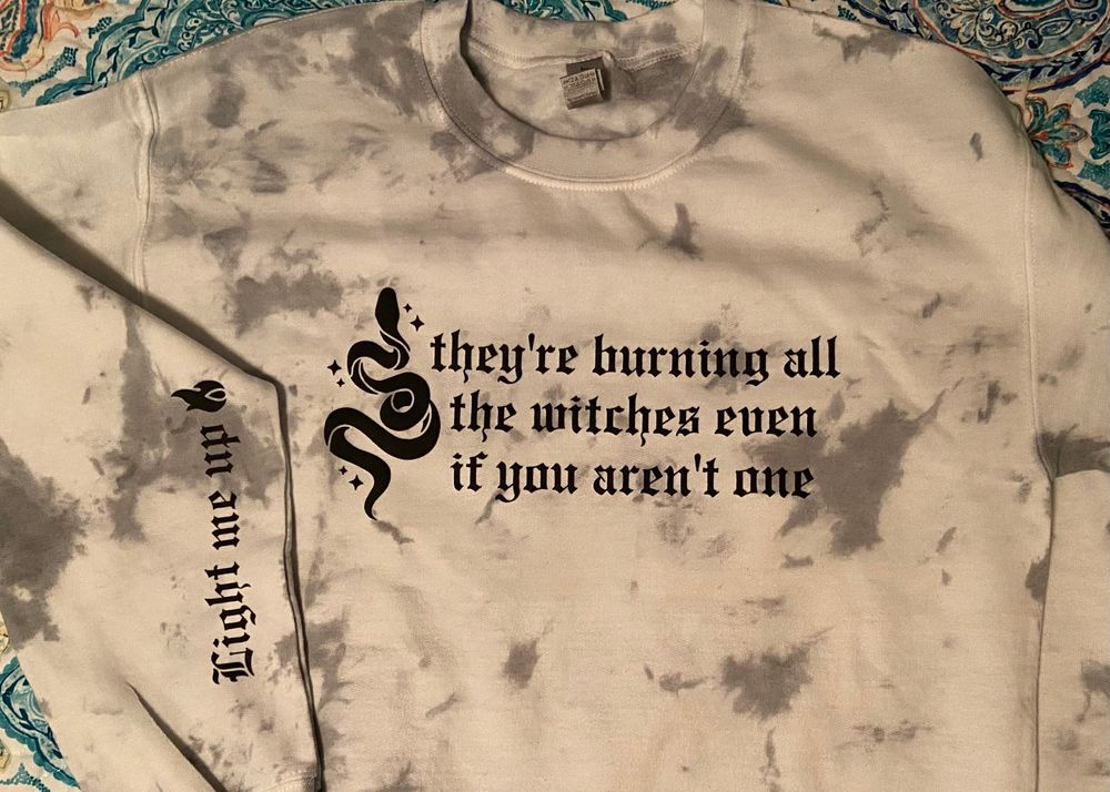 Gray and white tie die pullover sweater shirt that has an image of snake on it and the phrase “they’re burning all the witches even if you aren’t one” on the chest and “light me up” and a flame on the arm