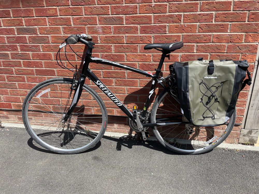 A black used Specialized Sirrus bicycle with green pannier bags, leaning against a brick wall.