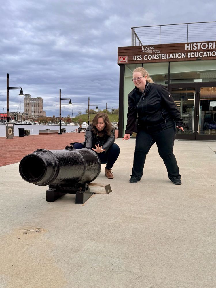 marisa & friend kailyn make dynamic goofy poses (marisa squatting with a determined face) behind the statue of a canon in baltimore's inner harbor