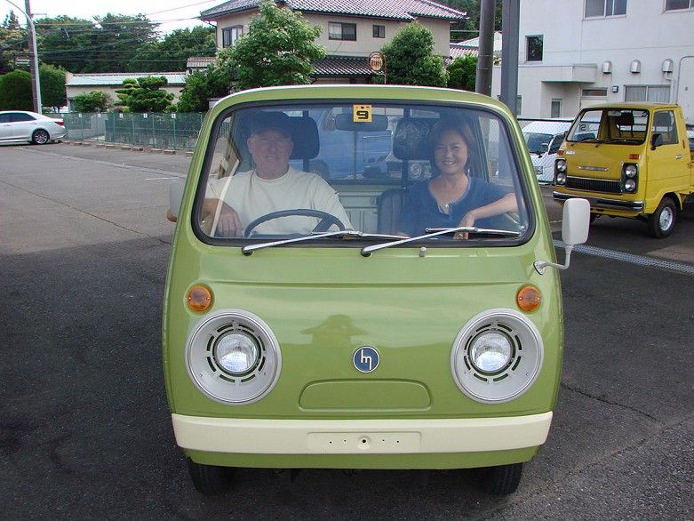 A green Mazda Porter kei truck, front on. There's a yellow kei truck with stacked headlights in back of shot that also looks fun.