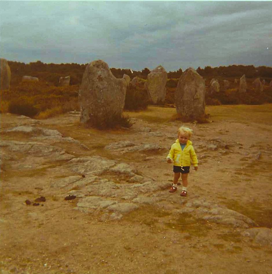 Picture of me aged perhaps 3 in a yellow windbreaker, blue shorts, and red shoes looking a bit windswept in front of standing stones perhaps somewhere in Brittany?