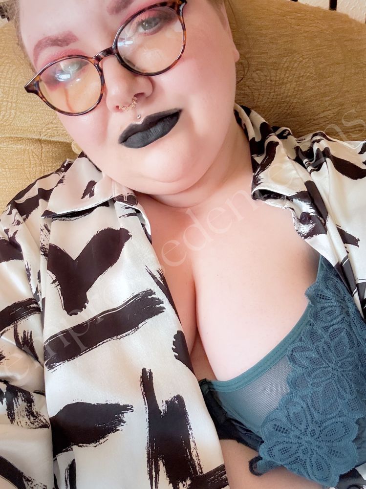A selfie. My left breast, in a green lacy bra, hangs out of my white blouse with black brushstrokes on it. My hair is tied up and I have my usual piercings and glasses. I have red eyeshadow and black lipstick on, and I smirk seductively at the camera