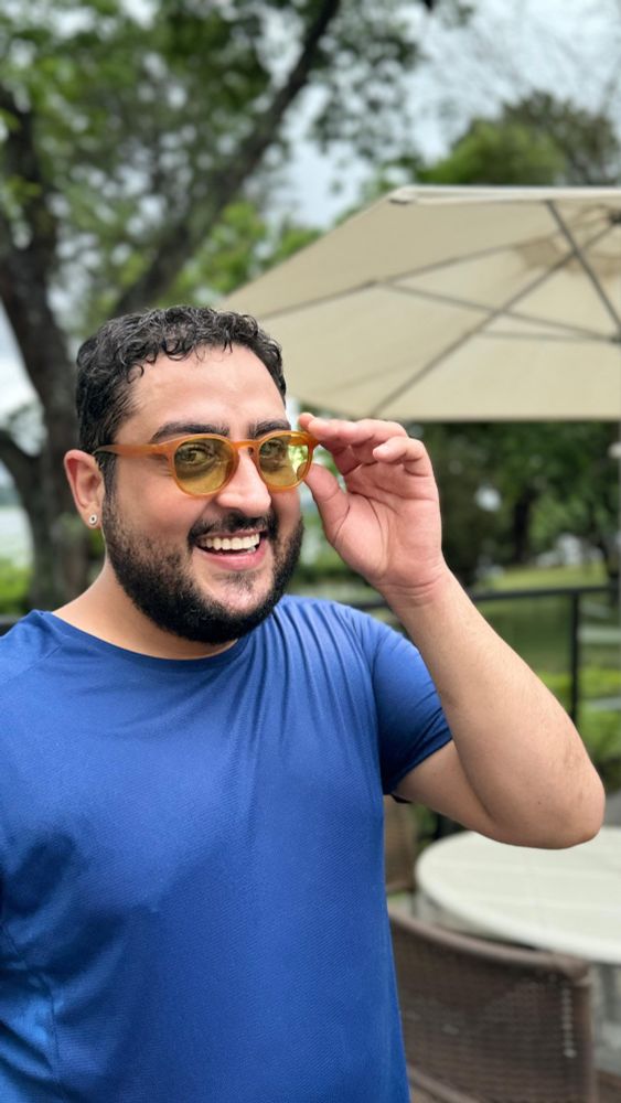 Homem de cabelos pretos, barba, camiseta azul e óculos de armação amarela sorri