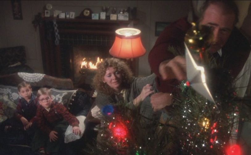 Still image from the film A Christmas Story. Ralphie, his mom, and his brother are watching his father precariously try to put the star atop their Christmas tree. The kids are sitting on a couch with a lit fireplace in the background. Their mom is trying to help and maybe keeping the dad from toppling over.