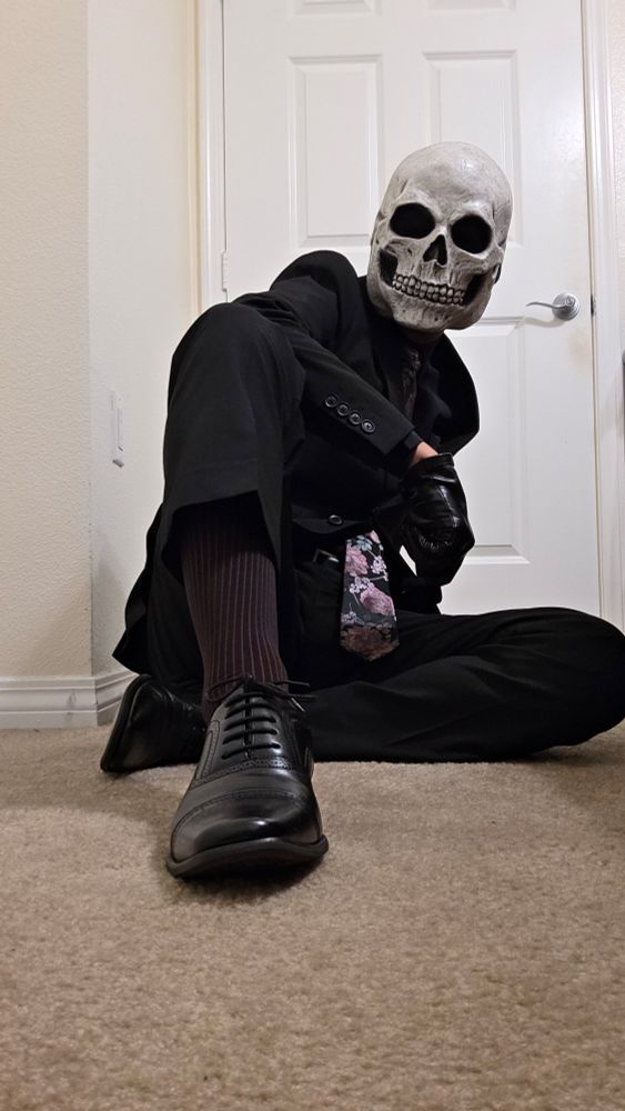 Skull head, black suit, black leather gloves, black dress shoes, pink floral tie. Sitting down.