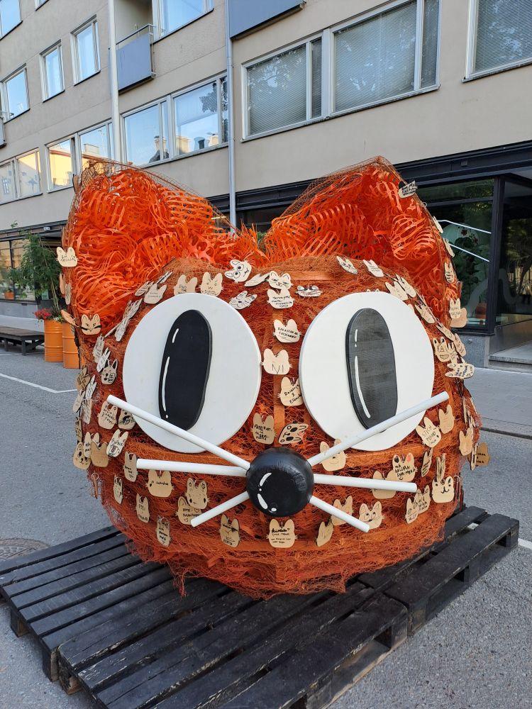 A stylized street-art sculpture of a cat head with large round eyes and orange hair.