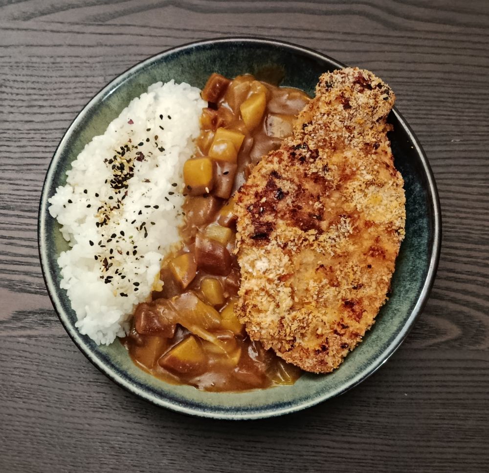 Assiette creuse grise en céramique. À gauche, du riz collant sur lequel a été saupoudré du furikake (mélange de sésame et algues séchées). À droite, une escalope de poulet panée. Au centre, du curry de légumes (oignons, carottes violettes, navet).