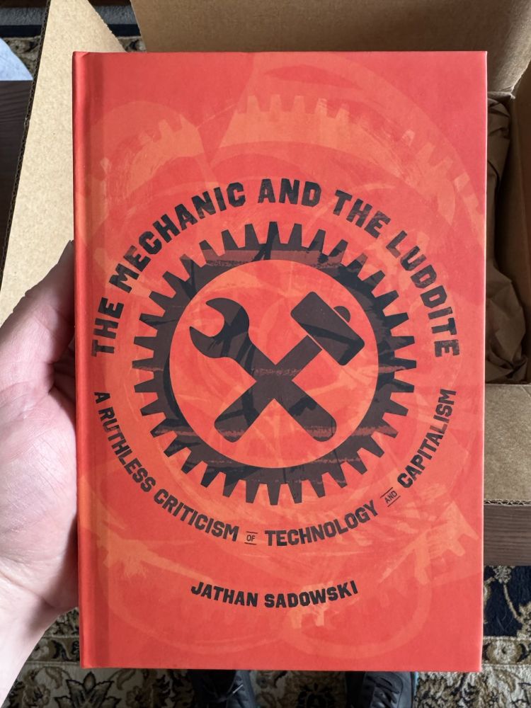 Photo of a white man's hand holding a book titled THE MECHANIC AND THE LUDDITE. subtitled A RUTHLESS CRITICISM of TECHNOLOGY and CAPITALISM. author JATHAN SADOWSKI. The cover is a mottled red and orange with an image of a circular toothed gear with a crossed wrench and hammer inside. The title is rockered over the top of the gear and the subtitle is rockered below the gear.
