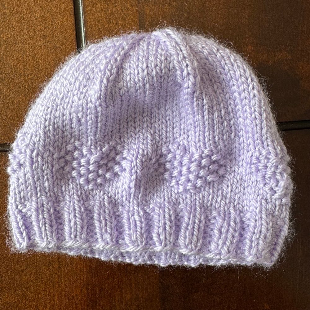 A baby-sized knit hat. The yarn is lavender in colour, and the side of the hat has hearts knitted into it.