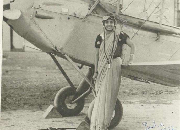 Sarla Thukral in B&W photo wearing a sari and flying helpmet with goggles standing in front of a 1930s plane