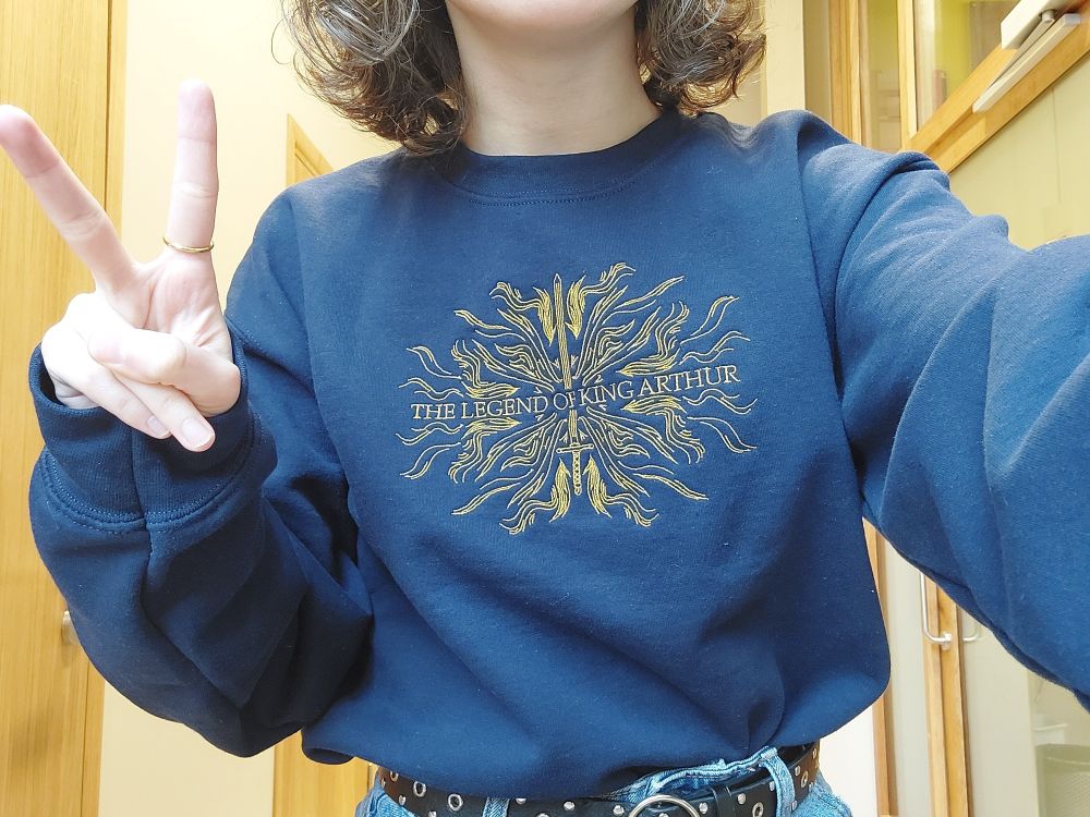 me wearing a navy sweater with "the legend of king arthur" and a sword embroided in gold on it. i'm also doing the peace sign. you can't see my face, just the top half of my body.
