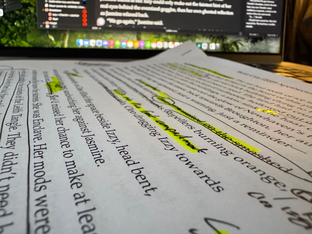 Close up of printed manuscript with handwritten & highlighted scribbles. Laptop screen in background. 