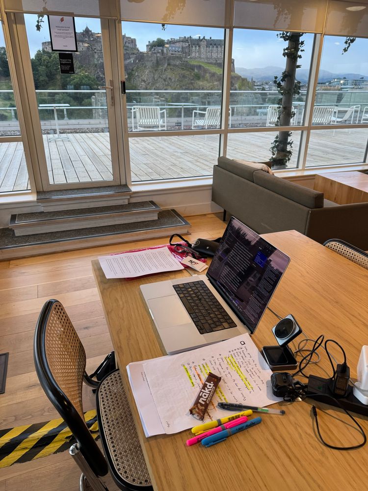 Office photo, laptop on wooden desk, writing notes, Edinburgh castle in background. 