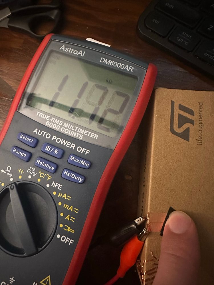 A red and grey multimeter showing 11 kohms with my finger on a black square of material, over two pieces of copper tape.