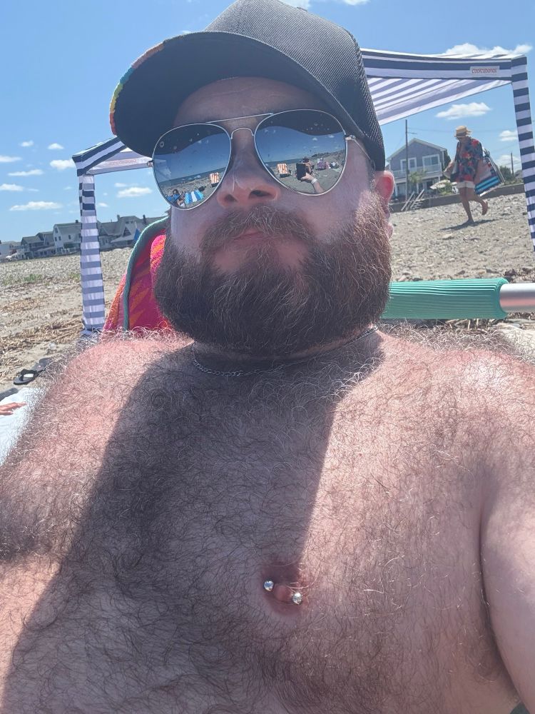 A selfie of me sitting on a sunny beach, in front of a blue and white striped canopy. I’m wearing a black ball cap with the pride colors embroidered on the edge of the brim, and a pair of silver mirrored aviators. The bright sunlight highlights the fur all over my chest and shoulders. 