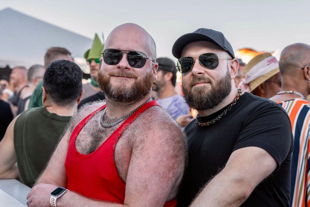A photo of me and my husband at tea dance in Ptown. I’m wearing a red stringer tank top that shows off my furry chest and shoulders, with a steel chain necklace and a pair of square black aviators. My husband is wearing a snug black T and a black ballcap, with a Byzantine link chain necklace in the bear pride colors, and a pair of black aviators with gold frames.