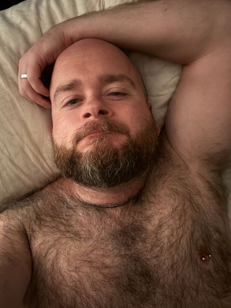 A closeup as I lie nude in bed, stoned and sleepy. My left arm rests on the pillow above my head, my hand curled around the right side of my head. My wedding ring catches the light. I’m bald and bearded with thick soft body hair that covers my chest and shoulders, and appears in a big tuft under my left arm.