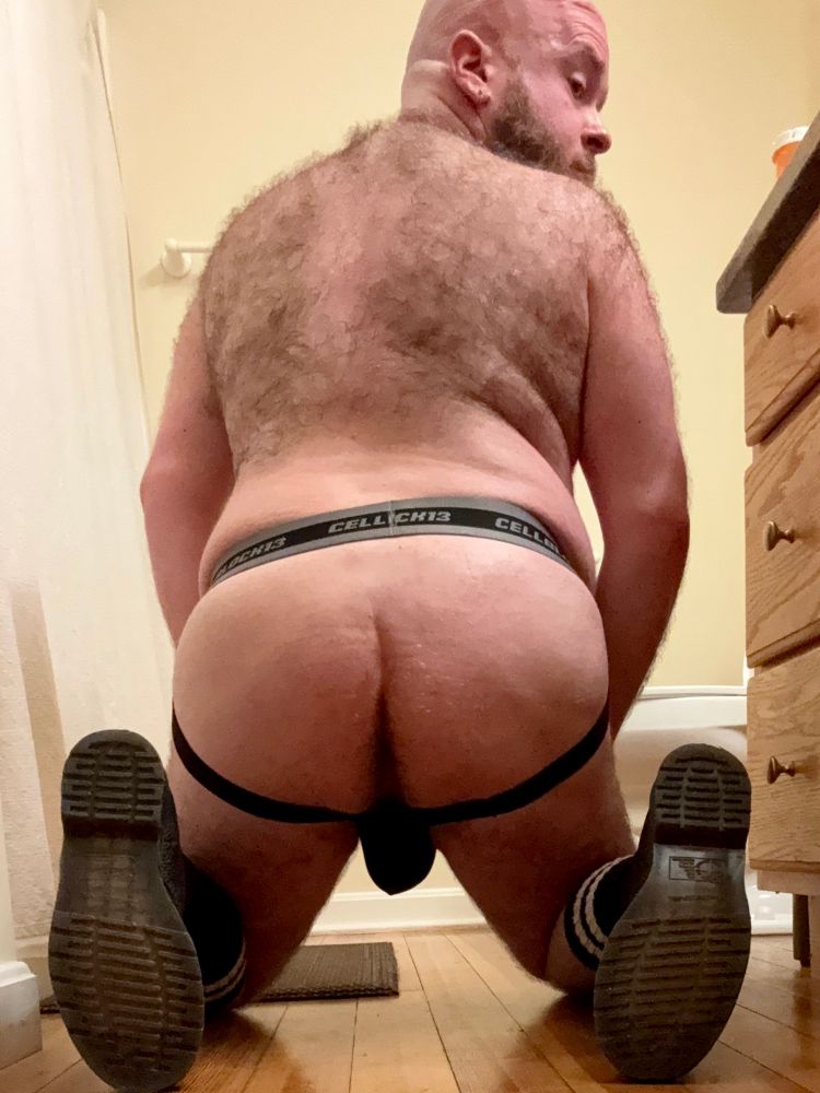 A photo of me from behind, kneeling on the bathroom floor. I’m wearing a black Cellblock 13 jockstrap, black leather boots, and black knee high socks with two white stripes around the top. I’m looking back at the camera over my hairy shoulder. My heavy balls fill the pouch of the jock, dangling between my thighs.